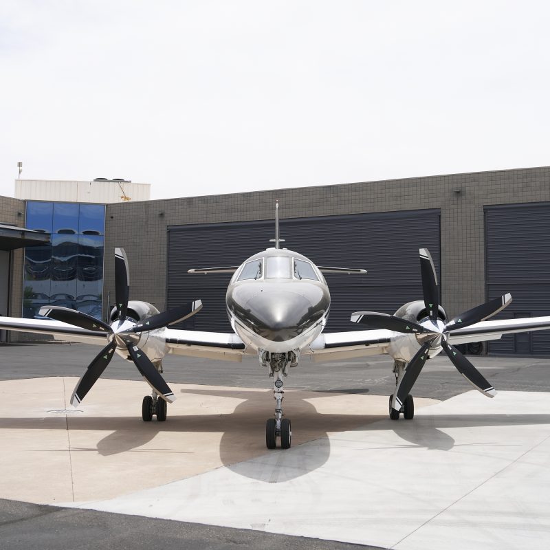 Front view of twin turboprop private plane with six-blade propellers parked on airport tarmac in front of hangar